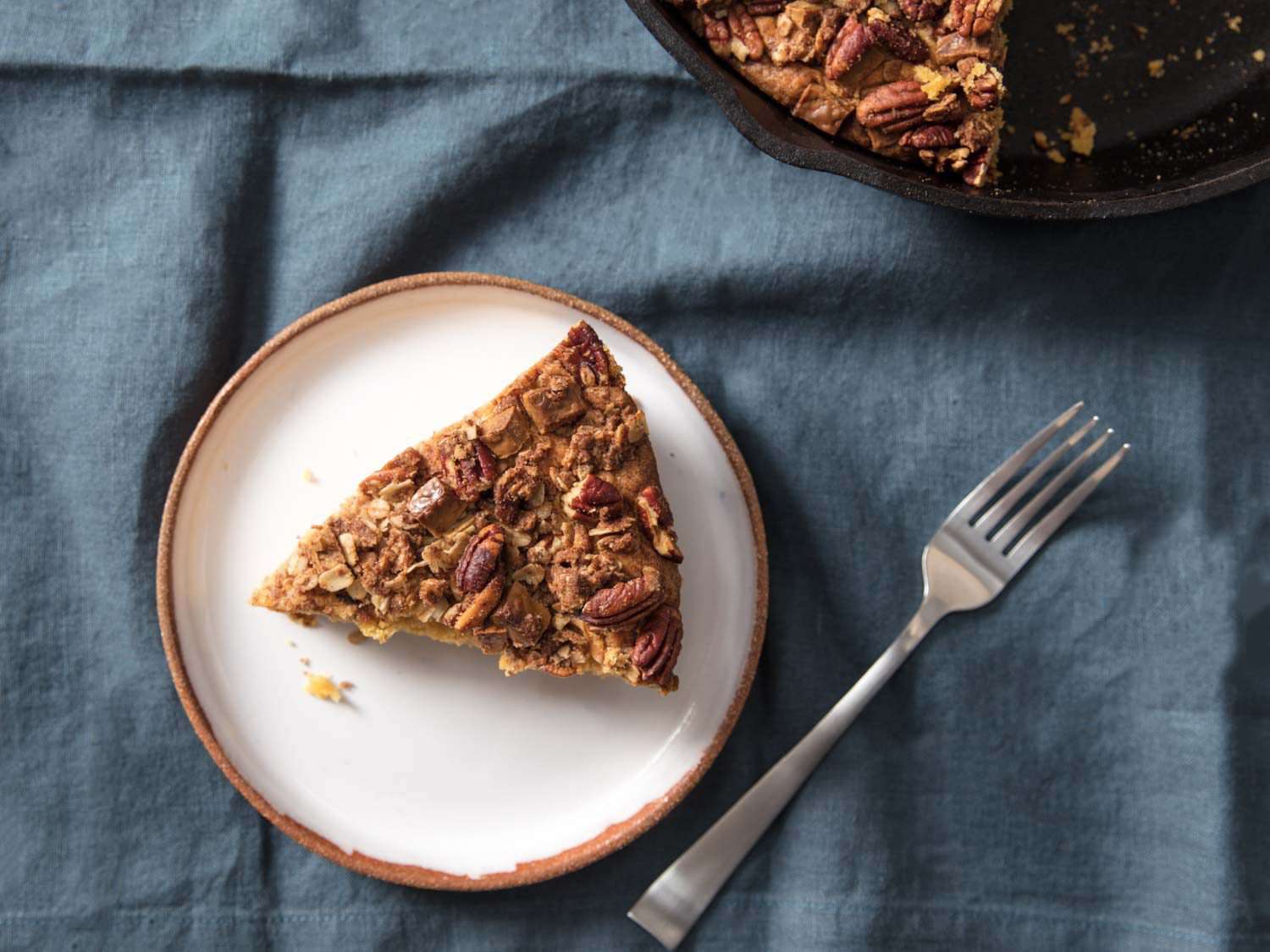 A slice of pumpkin streusel skillet coffee cake on a plate with a fork, on a blue tablecloth with the edge of the skillet cake in the corner. 