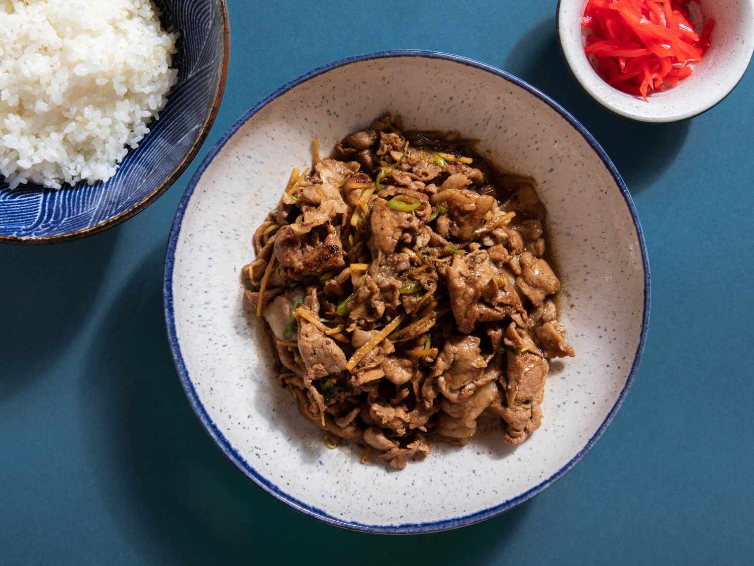 Cooked pork ginger in a serving bowl flanked by a bowl of white rice and ramekin of kizami shoga