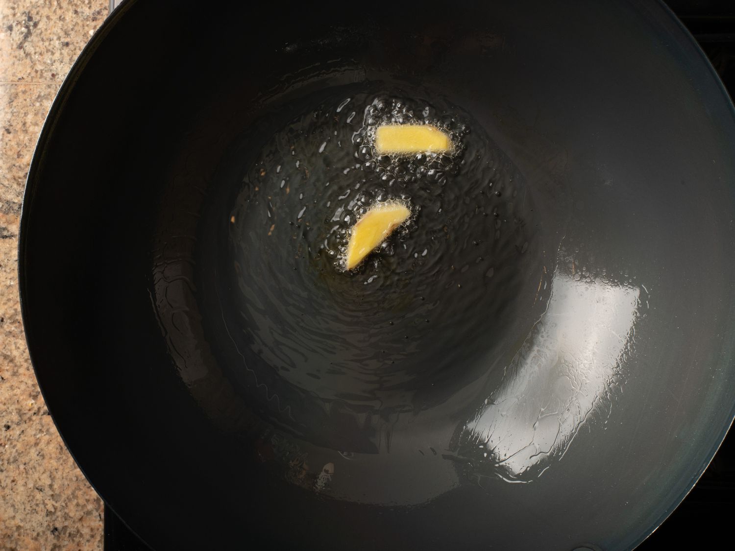 Two slices of ginger ginger in hot oil inside a black carbon steel wok. The ginger is surrounded by small bubbles, showing that the oil is hot.