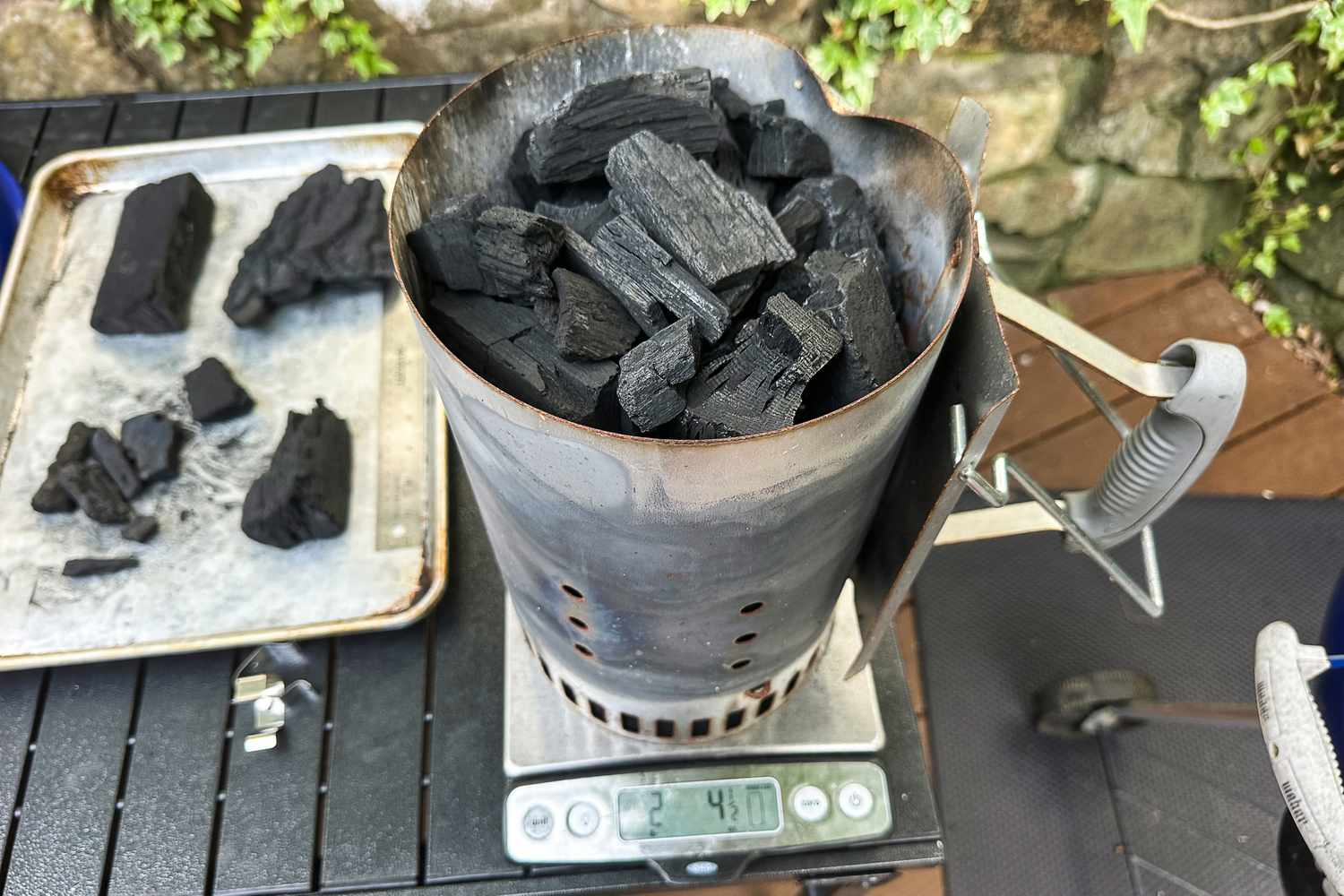 A metal chimney starter filled with pieces of charcoal placed on a digital scale with a tray of charcoal pieces nearby