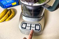 A person pressing the smoothie preset button on the Cuisinart Velocity Ultra 7.5 1 HP Blender with bananas and blueberries in the background