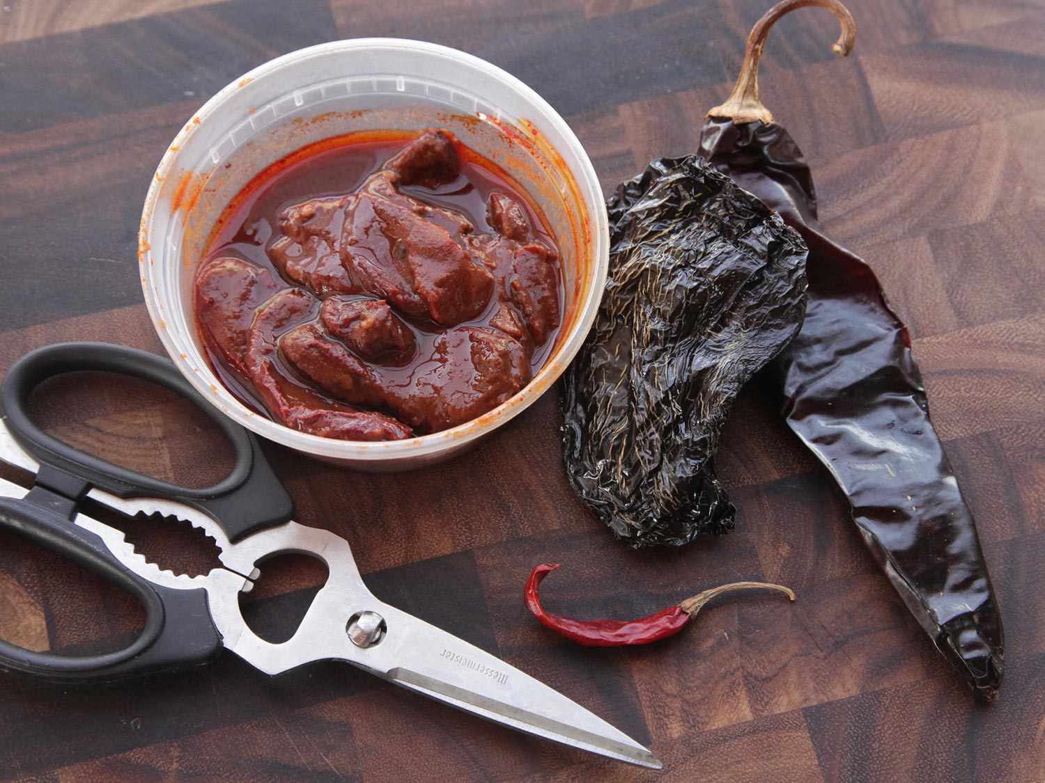 Several types of chiles displayed on a cutting board next to a pair of kitchen shears: a small deli container of chipotles en adobo and a dried ancho, a guajillo, and a chile de arbol.