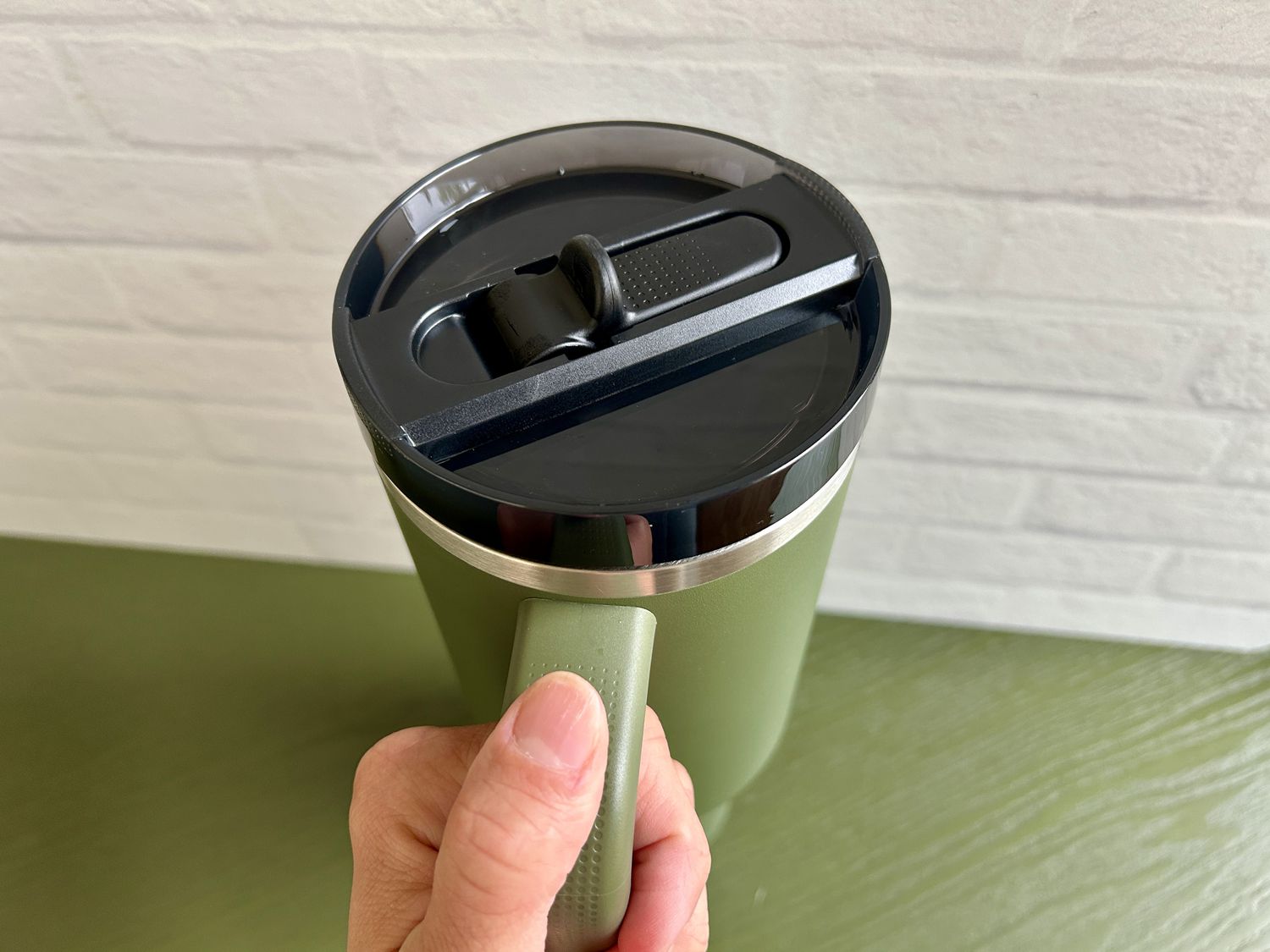 A hand holding a green Stanley Quencher ProTour tumbler in front of a white brick background