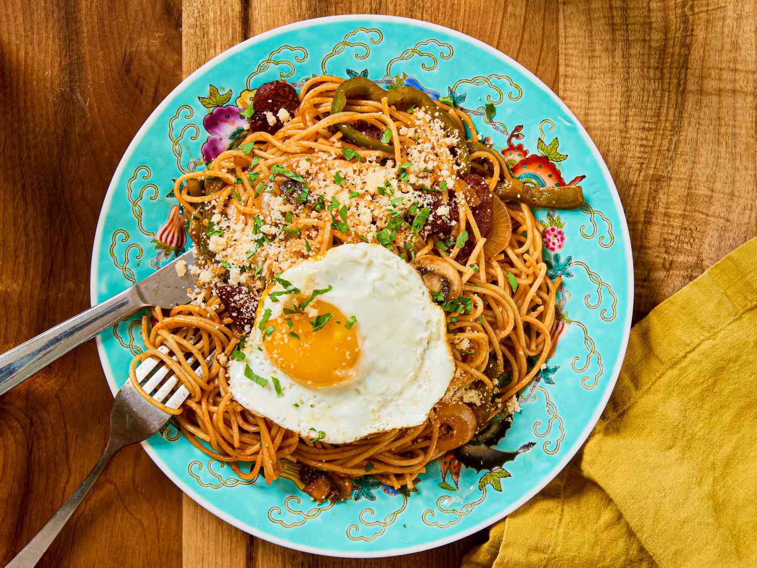 A plate of spaghetti Napolitan topped with a sunny-side-up egg, served on a decorative plate with utensils and a yellow napkin on the side
