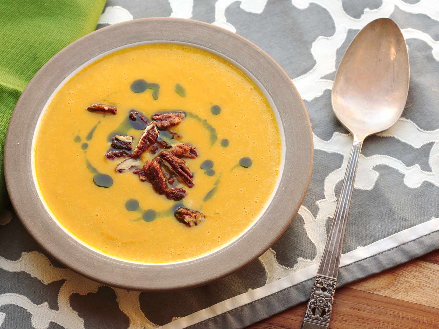An overhead view of a bowl of butternut squash soup, topped with a swirl of green sage oil and a handful of pecans, next to a spoon.