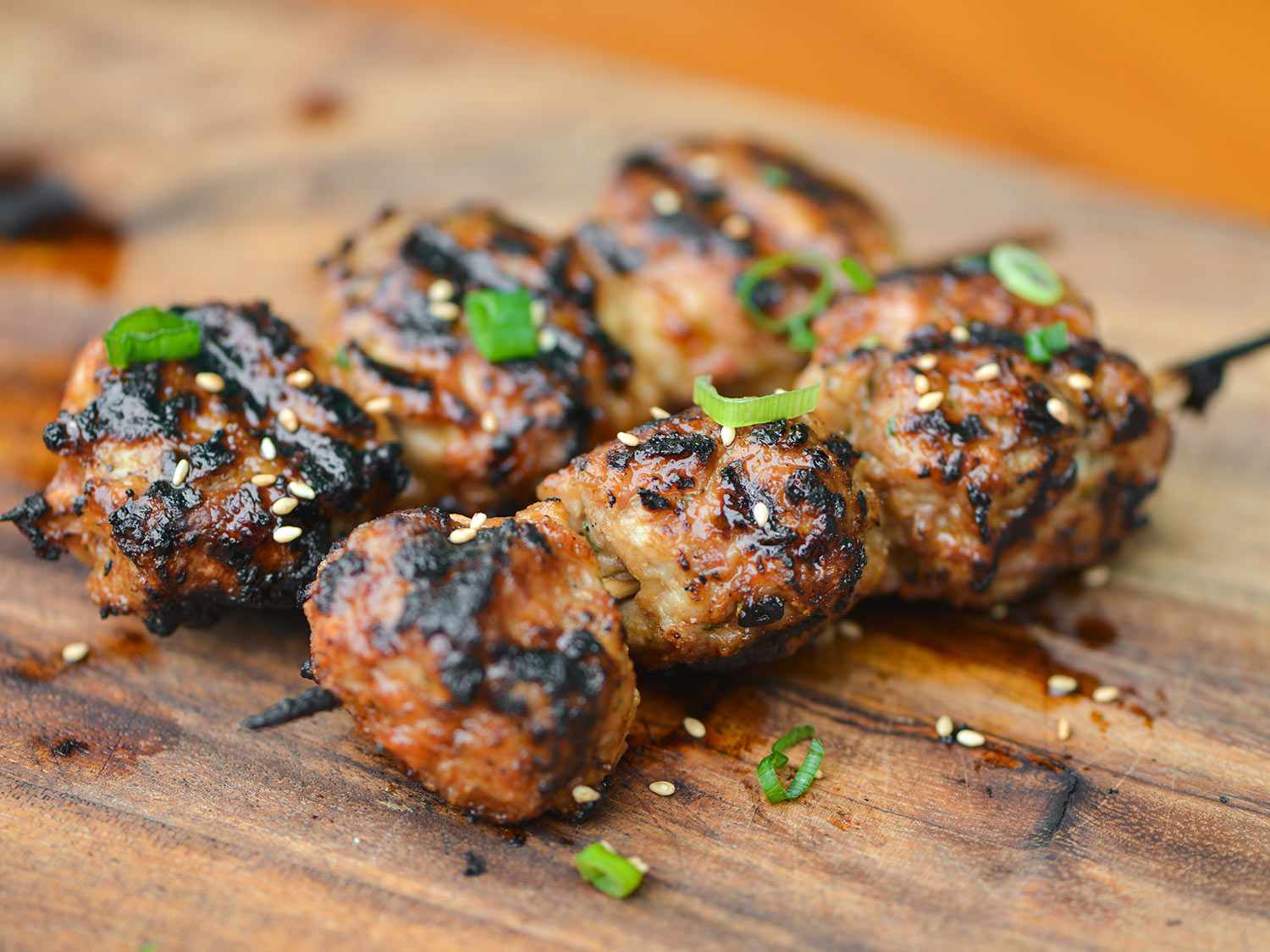Two skewers of Tsukune, Japanese chicken meatballs, resting on a cutting board and sprinkled with sliced green onions and sesame seeds 