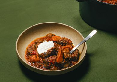Bowl of beef stew with vegetables and sour cream served with a spoon pot of stew in the background