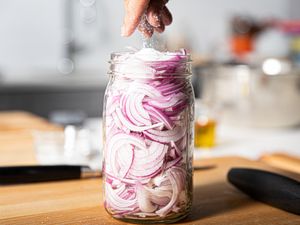 A hand adding liquid to a jar filled with sliced onions on a wooden countertop preparing quick pickled onions