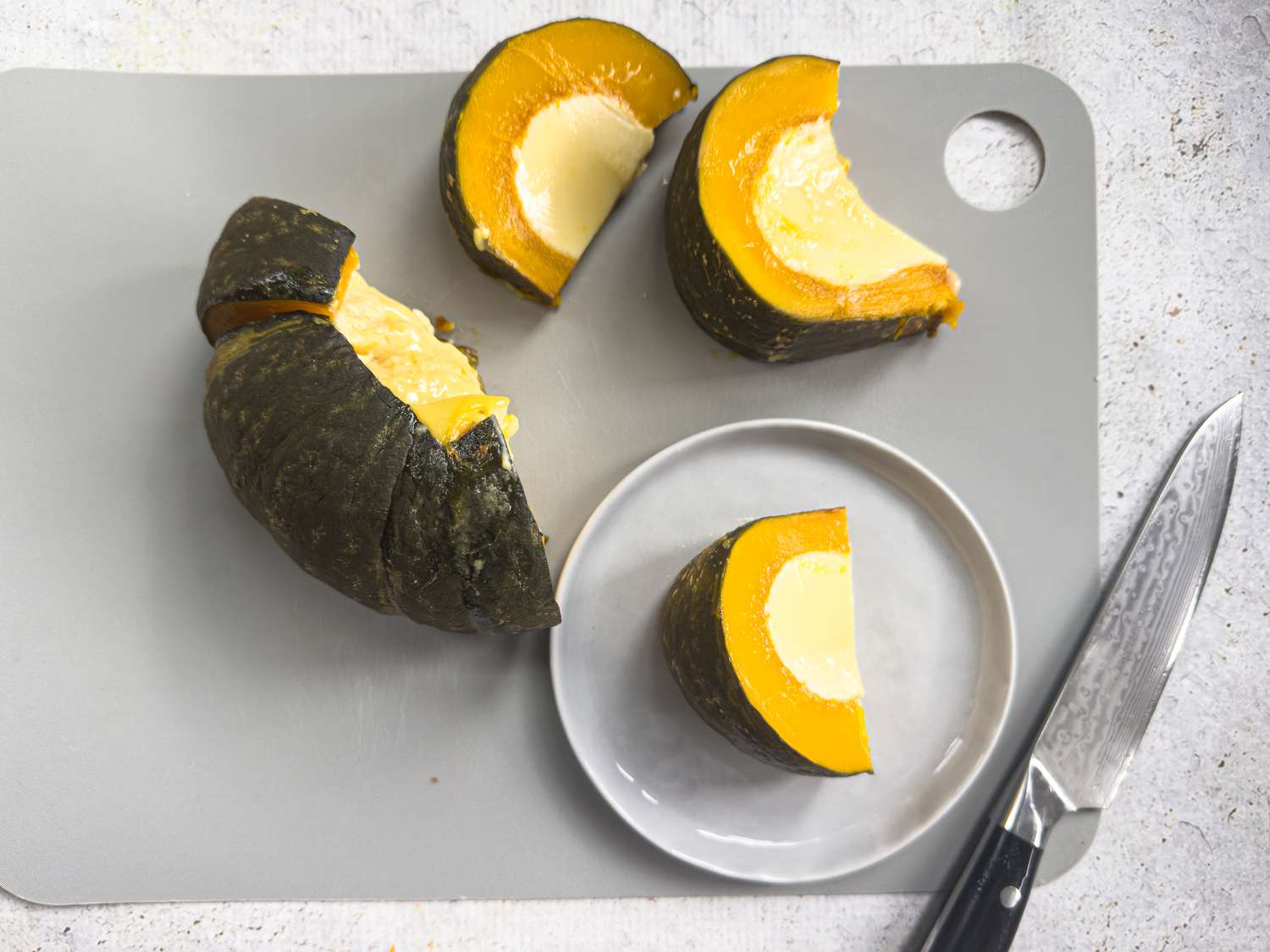 Sliced pumpkin custard. One slice on a grey plate, and the rest on a grey cutting board, with a knite on the edge of it, on a grey surface. 