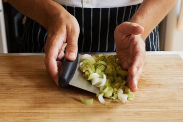 A person using a metal bench scraper to scoop up sliced leeks.