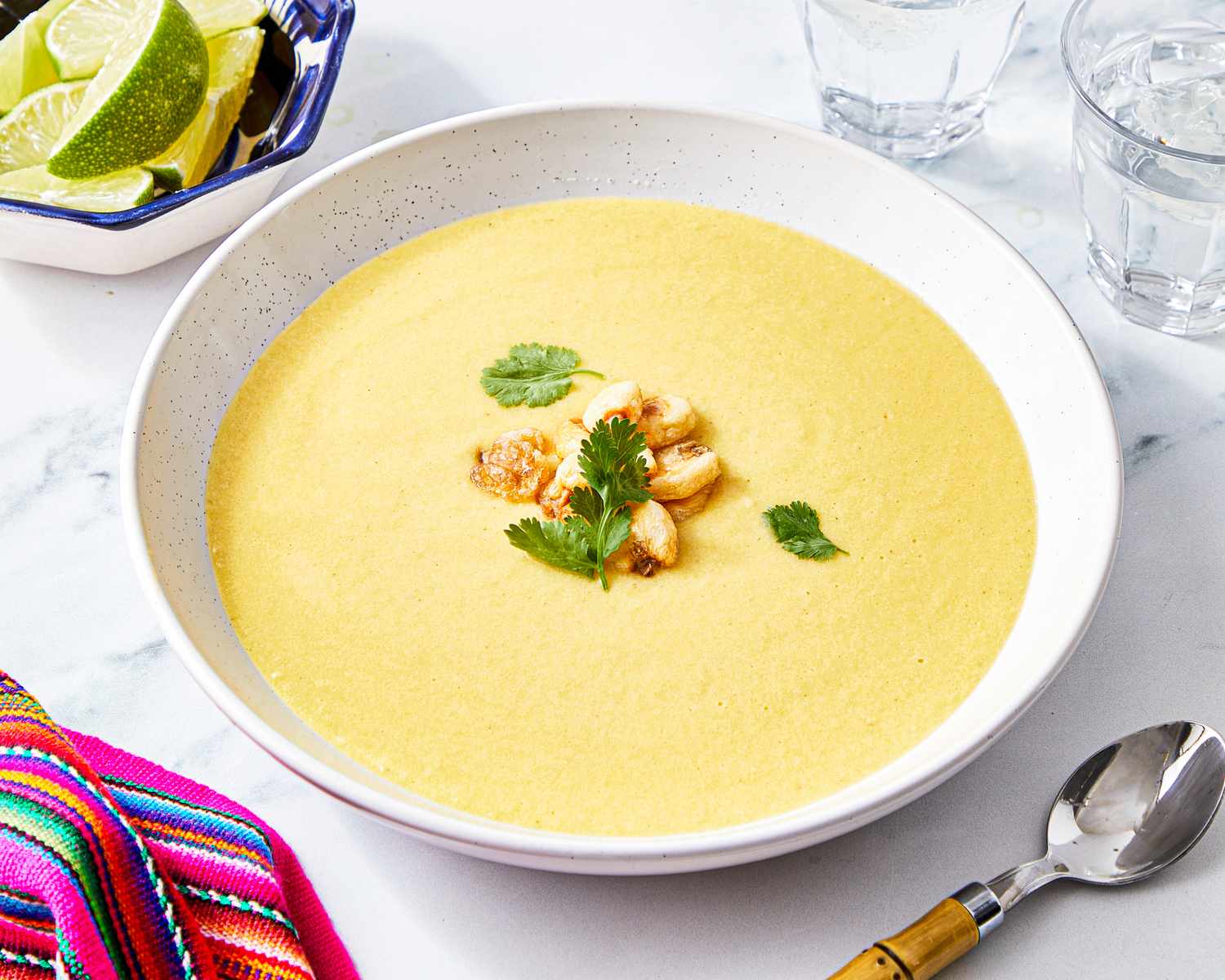 A bowl of corn soup garnished with herbs and nuts placed on a white countertop with a colorful fabric napkin and lime wedges nearby