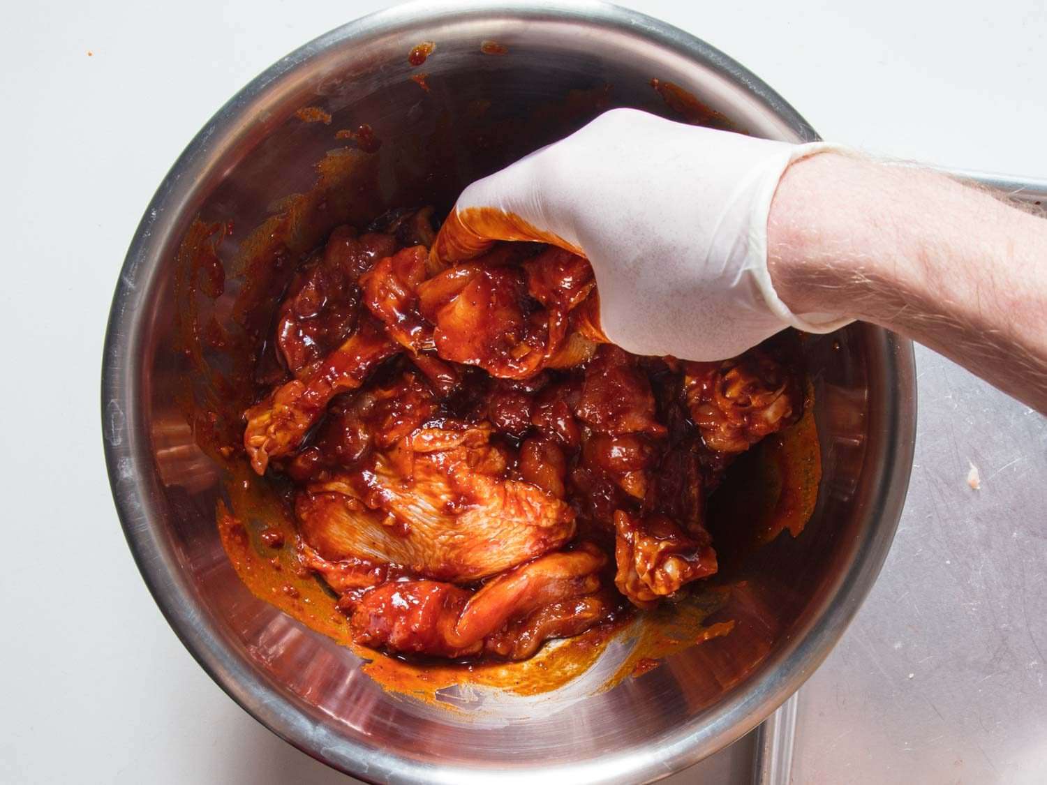 Massaging chicken pieces in red buldak sauce in a metal bowl.