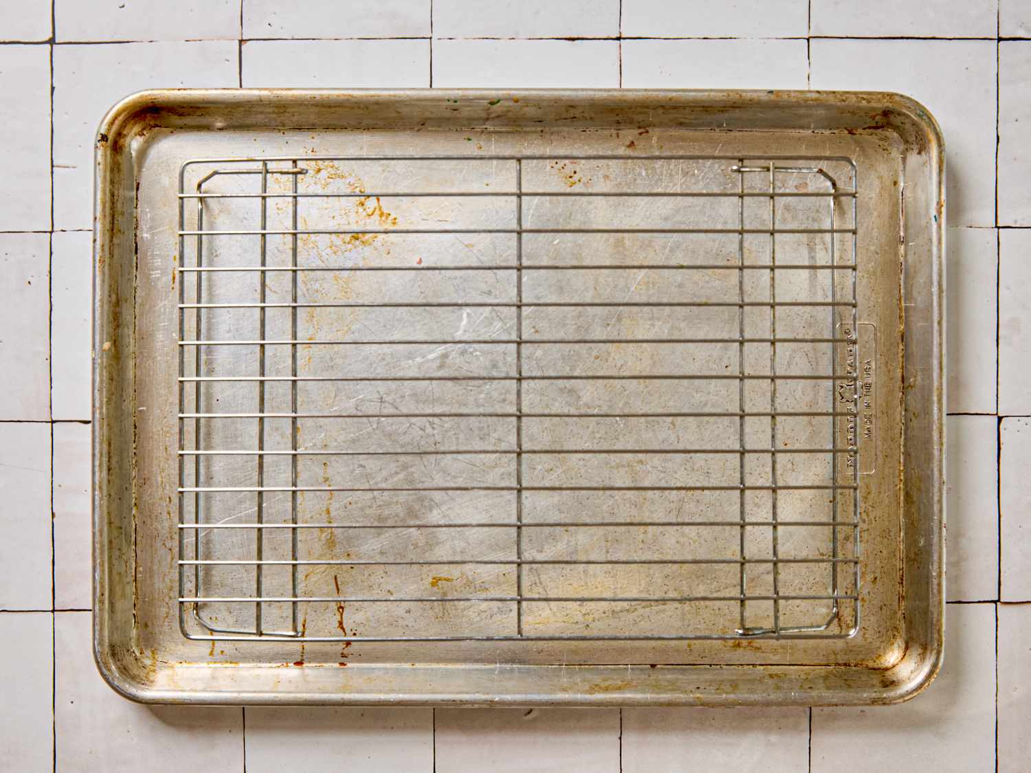 A used and slightly stained baking sheet with a metal cooling rack placed on it on a tiled surface