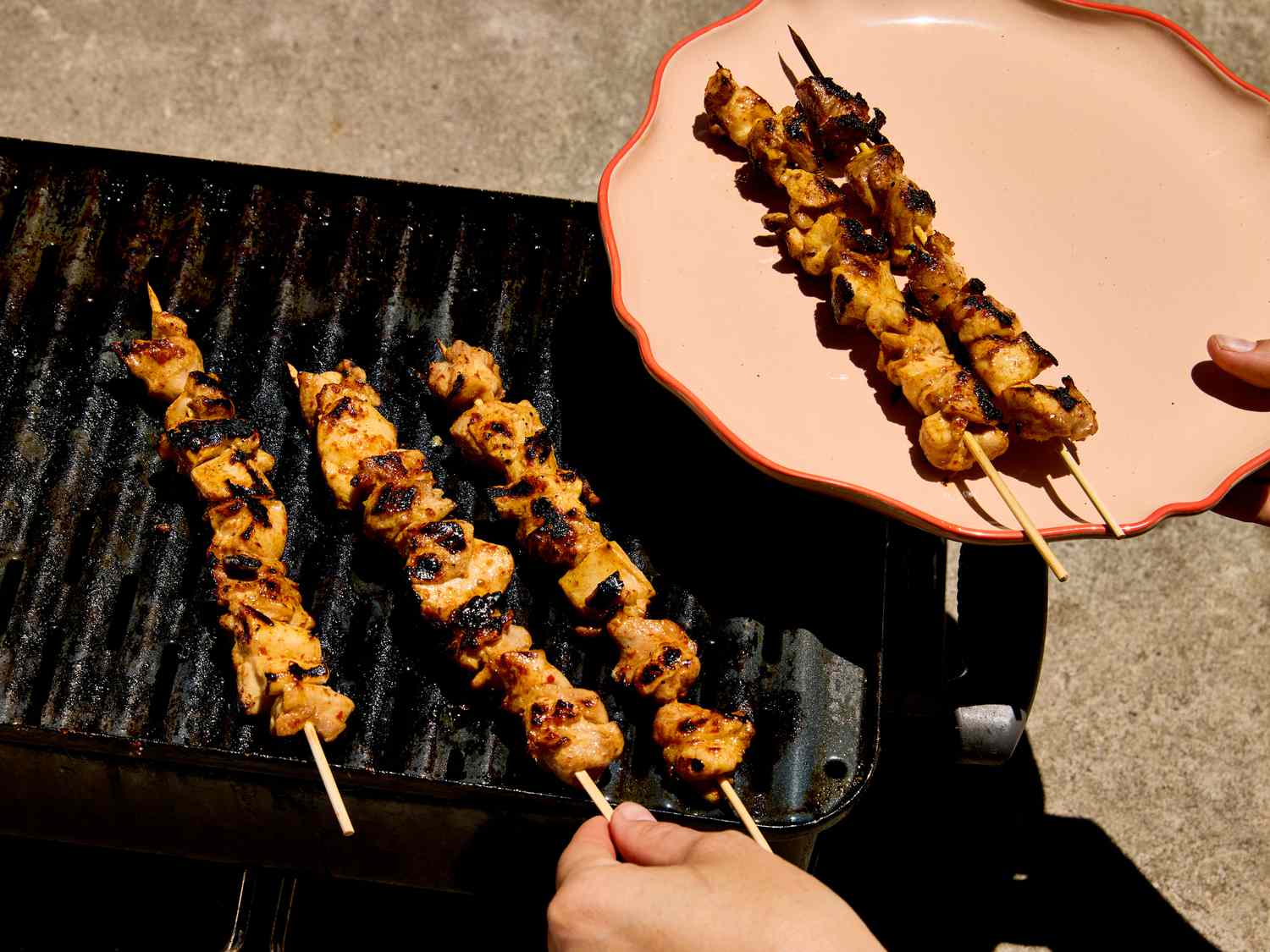 Grilled chicken skewers being transferred from the grill to a plate by a person
