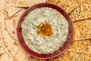 A plate of creamy spinach dip garnished with fried onions surrounded by flatbread and sesame crackers