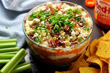 7-layer buffalo chicken dip, served in a glass bowl with tortilla chips and celery sticks.