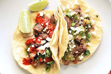 Overhead shot of two tacos de lengua on a plate. One is garnished with a red salsa and the other sports green salsa. Both are sprinkled with queso fresco, scallions, and cilantro. Two lime wedges flank the tacos.