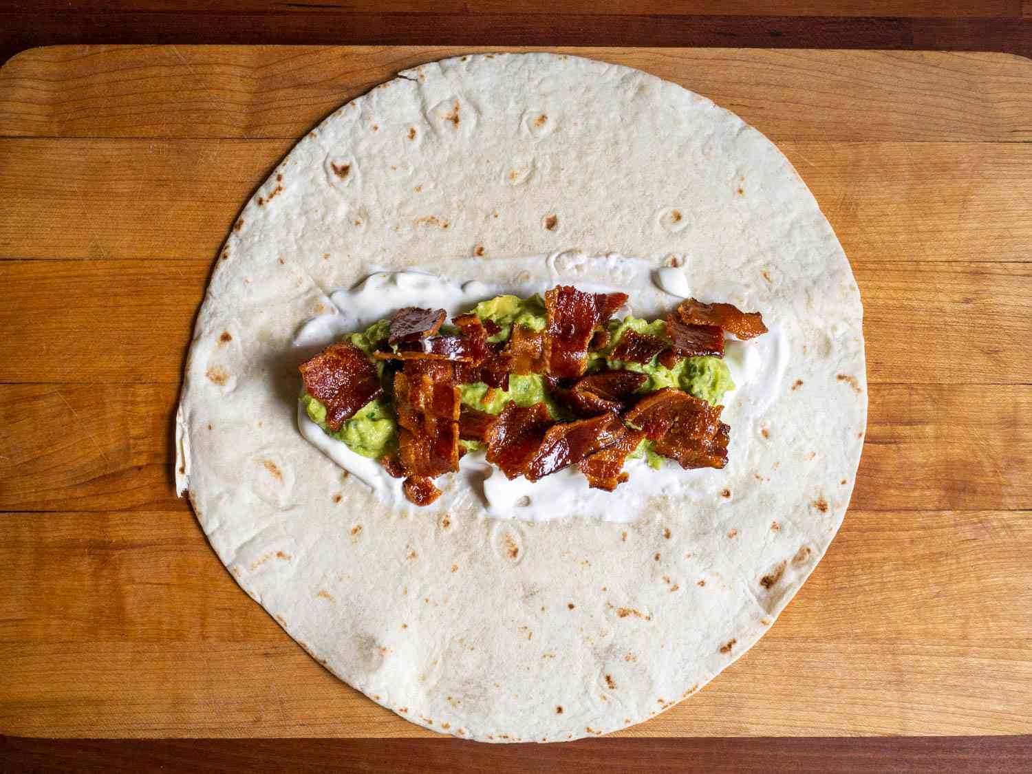 Overhead view of the burrito being assembled. Sour cream, guacamole, and bacon have been added to the center of the tortilla.