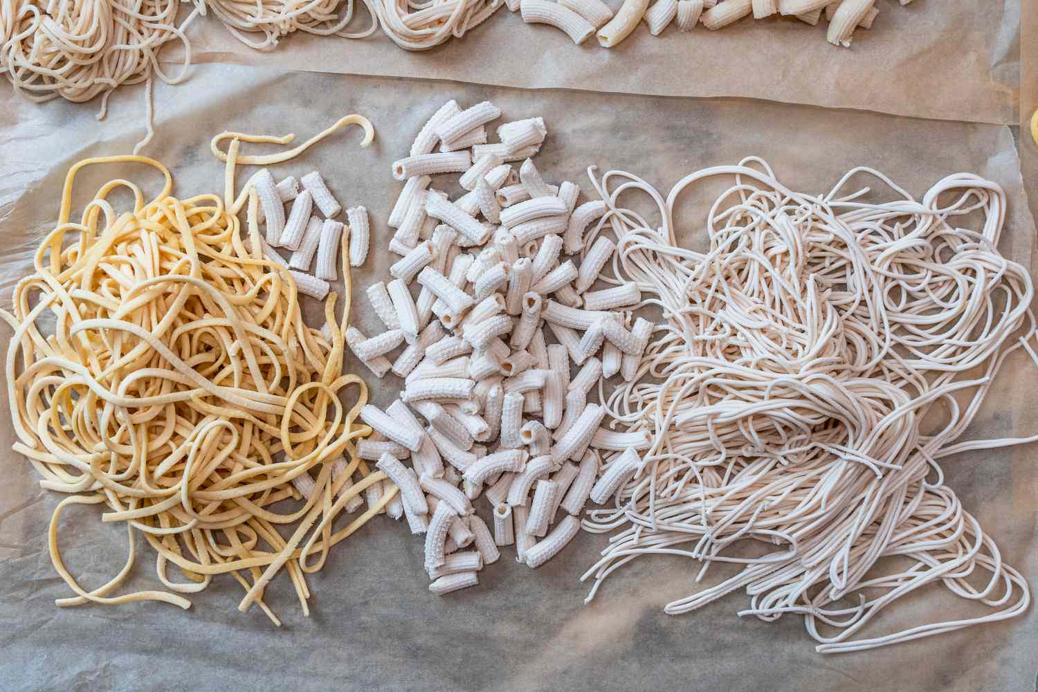 Three piles of spaghetti, fettuccine, and penne on a piece of parchment paper.