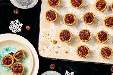 Decorative board with pican tassies on it, on top of a dark green velvet fabric. Smaller plate with 3 tassies on it, one bitten into. 2 cups of water.