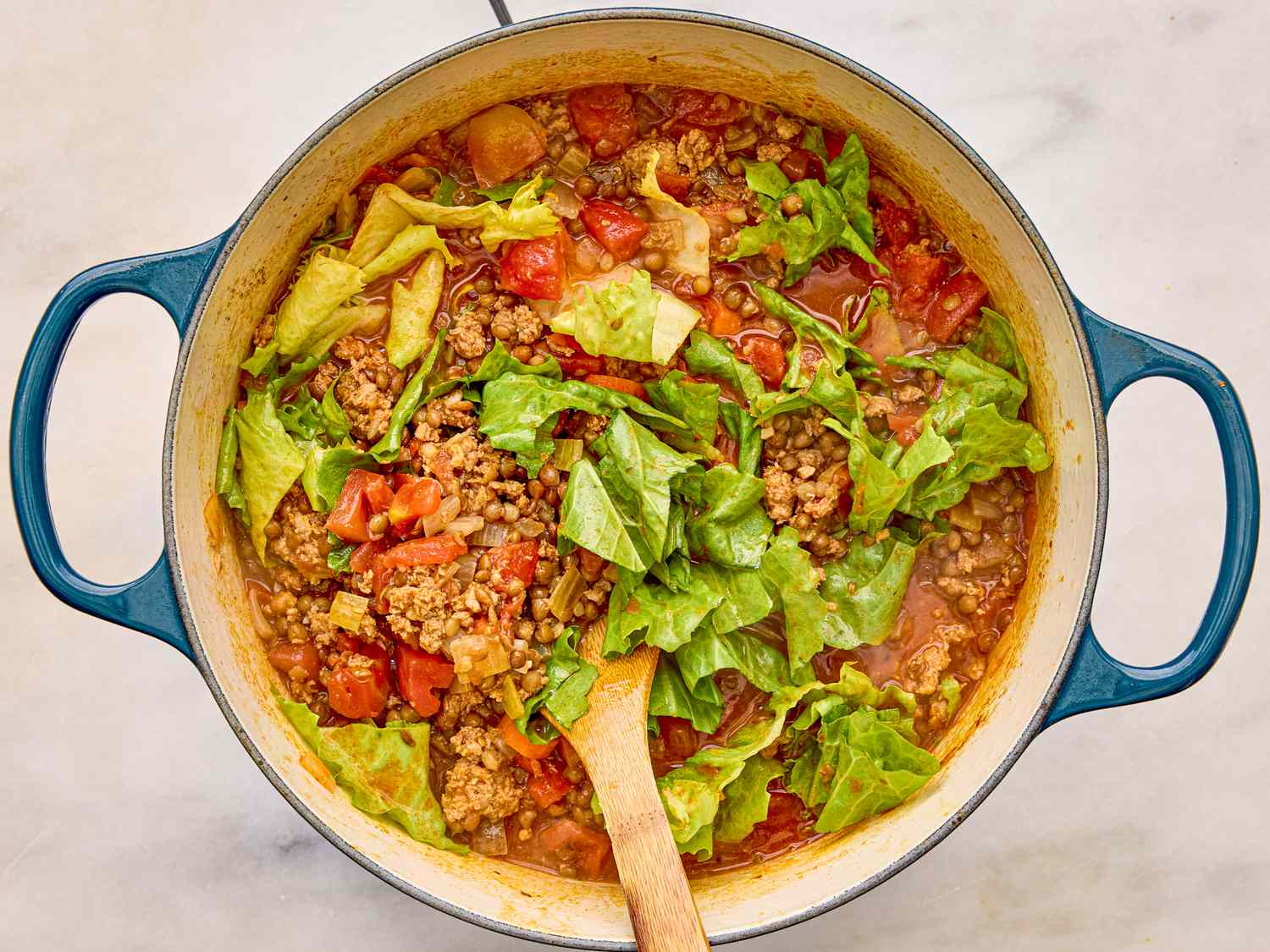 A pot of sausage and lentil soup with chopped greens and a wooden spoon