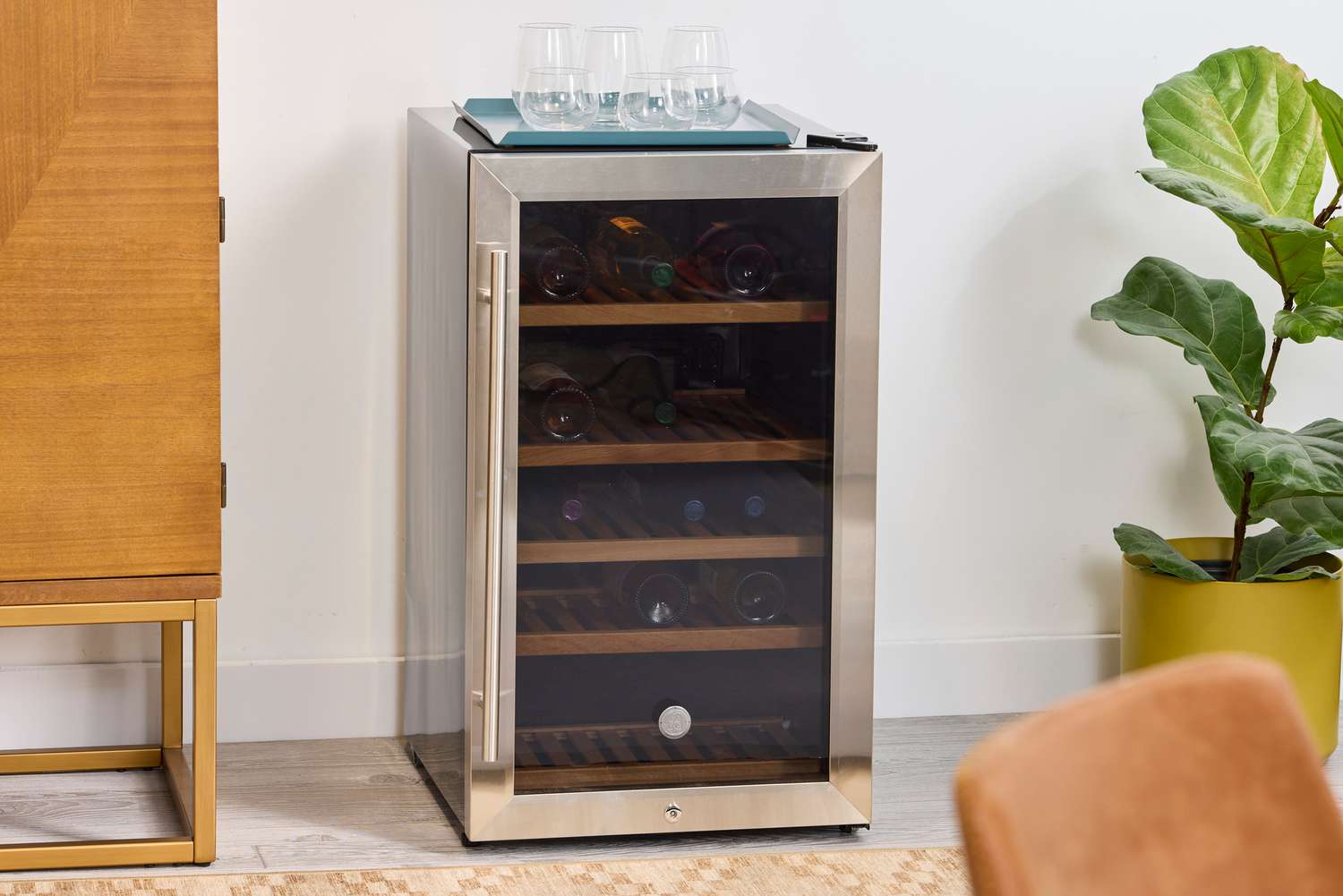 Compact wine refrigerator with glass door, bottles inside, glasses on top, wooden cabinet and indoor potted plant nearby