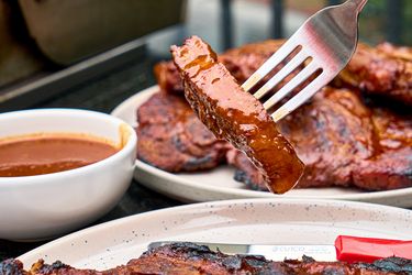A fork holding a piece of cooked pork steak near a dish of sauce with a background of additional steaks on plates