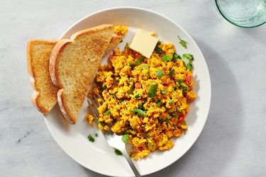 The anda bhurji on a white ceramic plate with toasted white bread and a pat of butter on a stone counter 