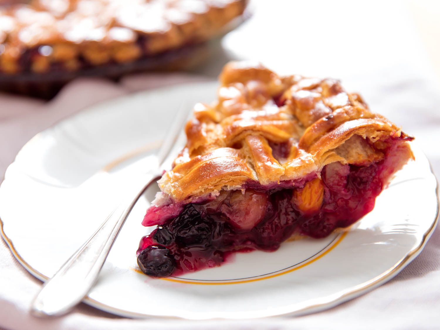 A slice of mixed-fruit pie with a golden basketweave-style top crust. The filling looks chunky, with tender, pitted cherries and whole blueberries suspended in a vibrant, jammy filling. 