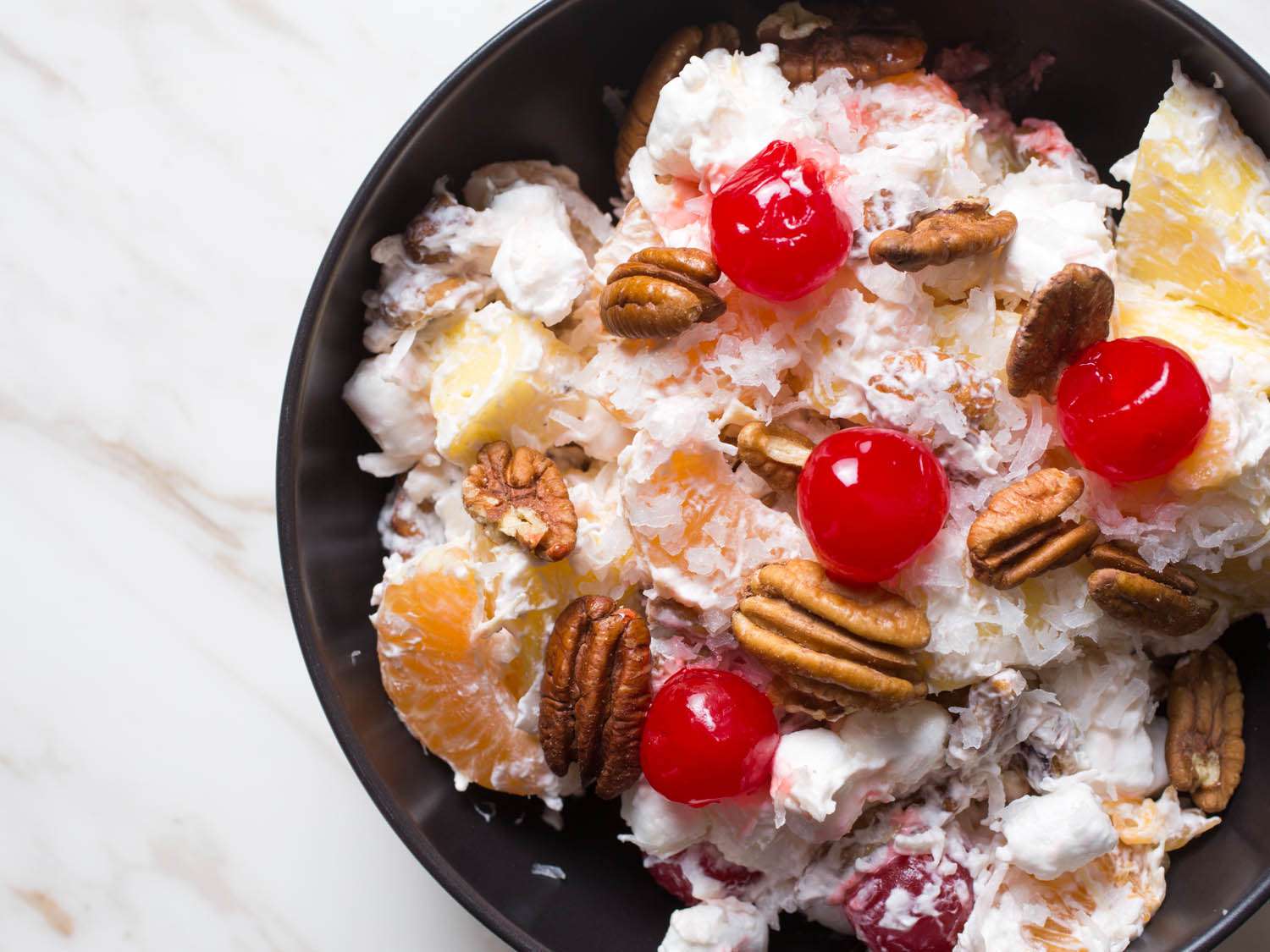 A bowl of ambrosia, a fruit salad with coconut, pineapple, pecans, orange segments, mini-marshmallows, and cherries.