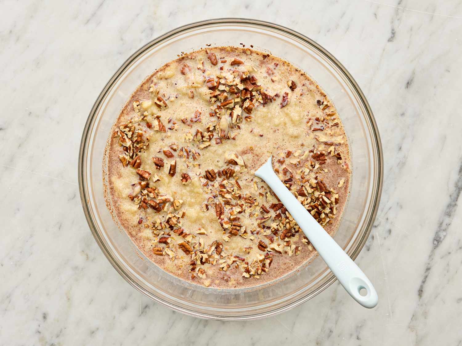 A dish of banana bread pudding topped with chopped nuts and a white serving spoon placed inside on a marble surface