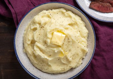 A bowl of fluffy mashed potatoes sits on a table. It's topped with a pat of melting butter and a grinding of fresh black pepper.