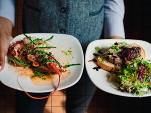 A waiter presenting two plated gourmet dishes one featuring seafood and the other a salad with toast