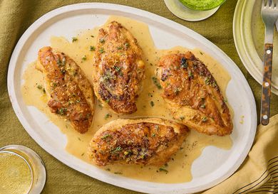 Lemon Pepper chicken on a white platter on a mustard colored textile, with yellow napkin, plates and drinks surrounding it