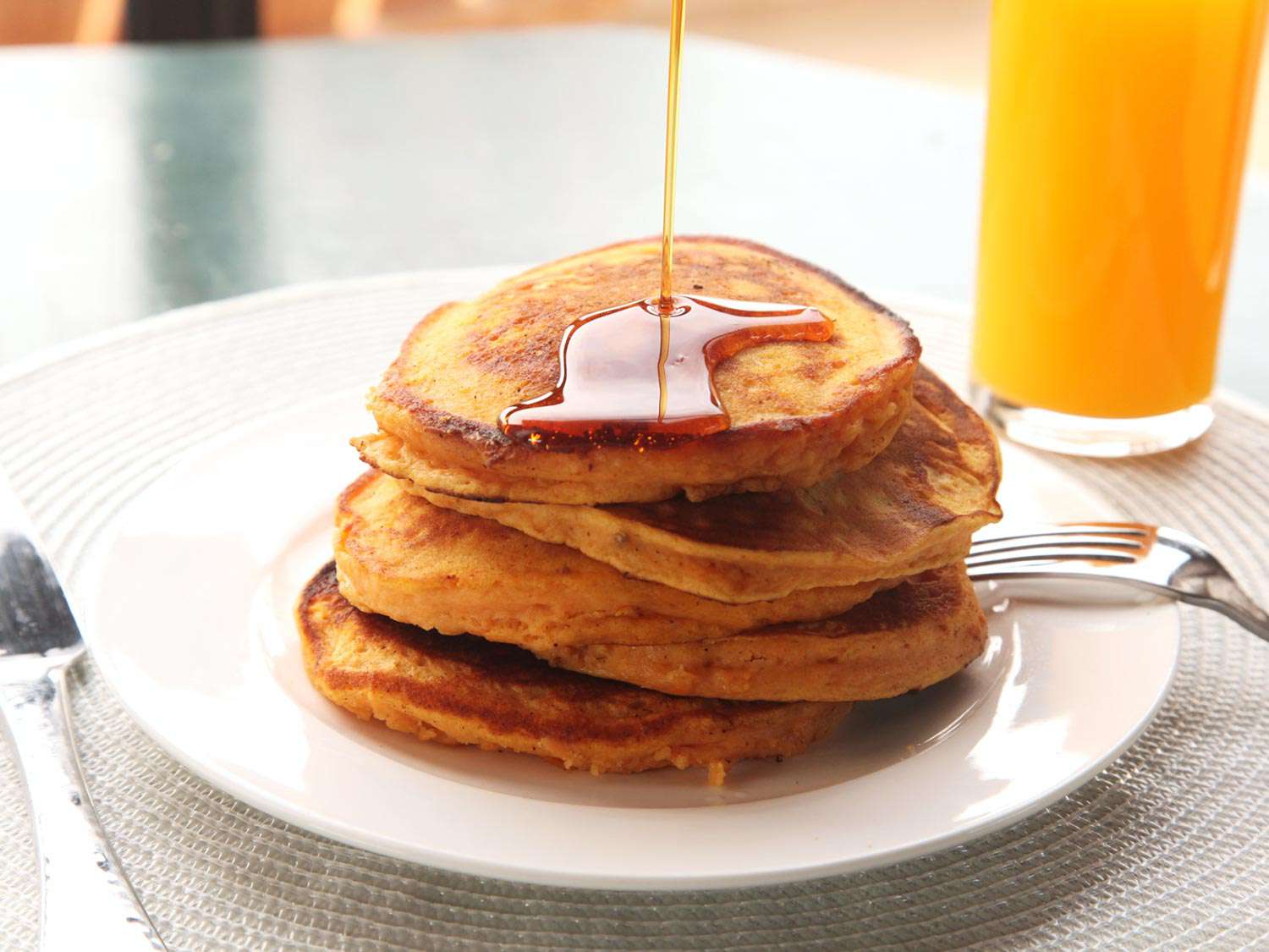 Maple syrup being poured over a stack of sweet potato pancakes on a white plate with a fork next to a glass of orange juice.