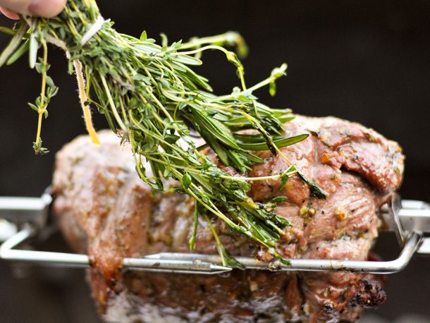  A boneless leg of lamb on a rotisserie that is being basted with a bundle of fresh herbs.