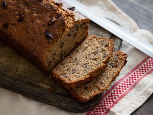 Banana bread with pecans sliced on a small wooden cutting board on top of a white kitchen towel with knife on the side.