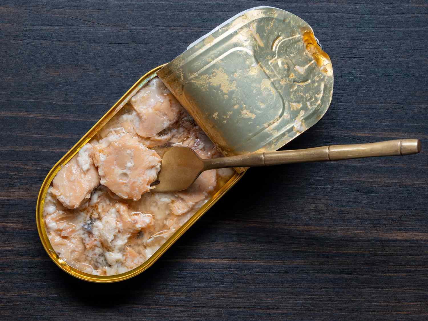 An open tin can with fish chunks in sauce on a wooden surface a fork resting inside the can