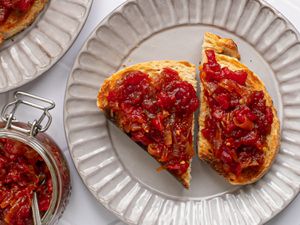 Two slices of bread topped with tomato and onion jam on a ridged cream colored ceramic plate. In the left corner of the image is a glass jar filled with more jam, and in the top left corner is a portion of another plate with more toast on it.