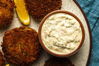 A small bowl of extra tangy tartar sauce on a speckled ceramic plate. The tartar sauce has crab cakes and lemon wedges around it.