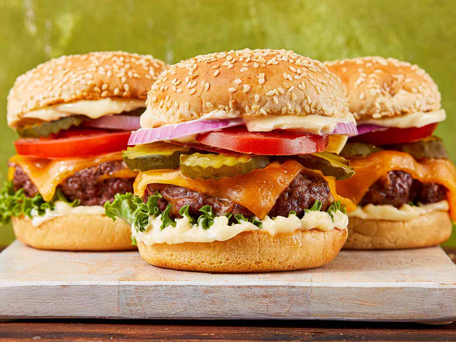 Three cheeseburgers with lettuce, tomato, pickle, onion, and cheddar cheese on a sesame seed bun displayed on a cutting board