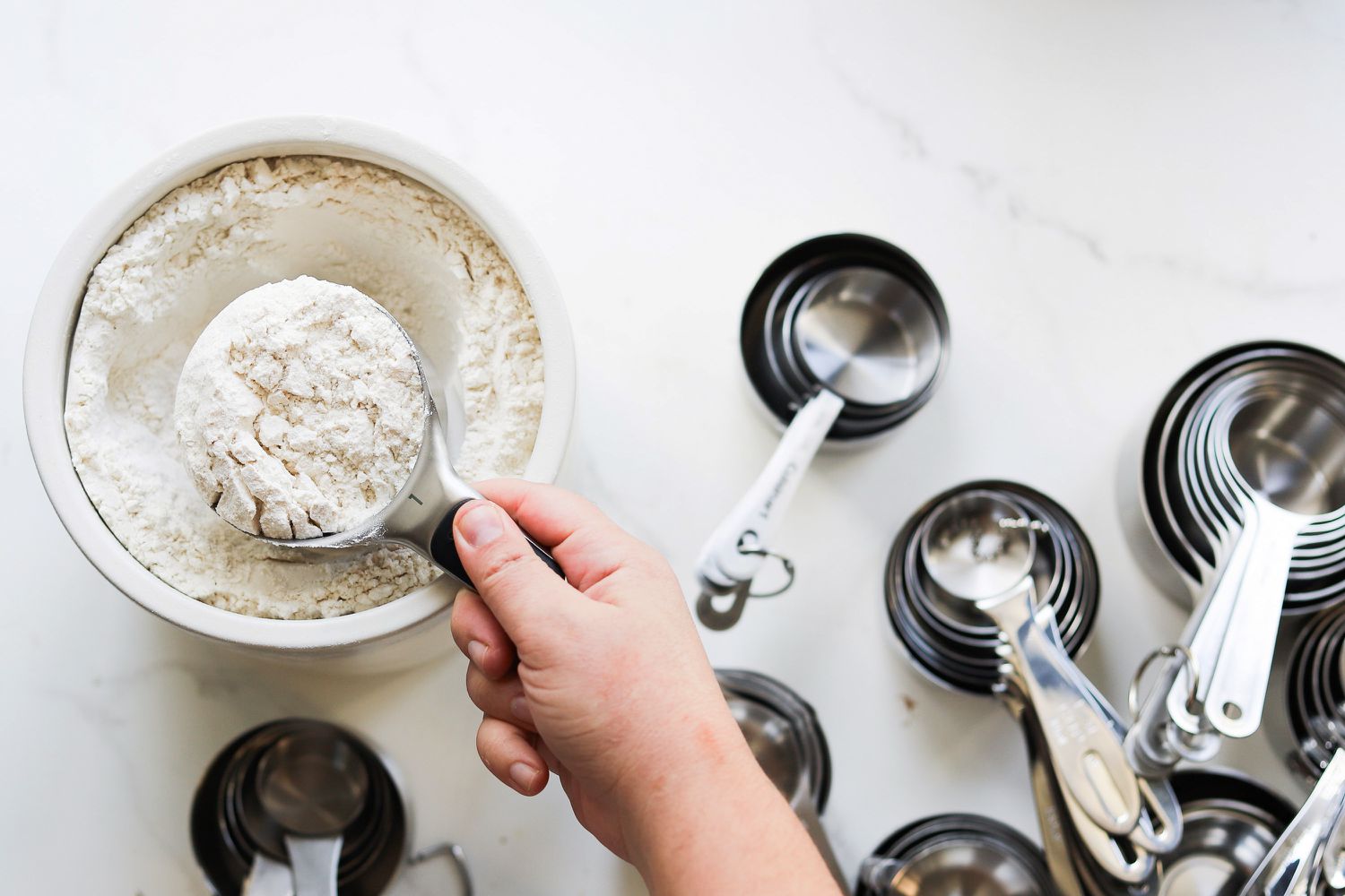 Scooping flour from a bowl using the OXO Stainless Steel Measuring Cups surrounded by stacks of measuring cups