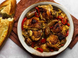 Chicken scarpariello in a round ceramic platter. The platter is on a maroon colored piece of cloth, and there is a broken loaf of bread on the left hand side of the image.