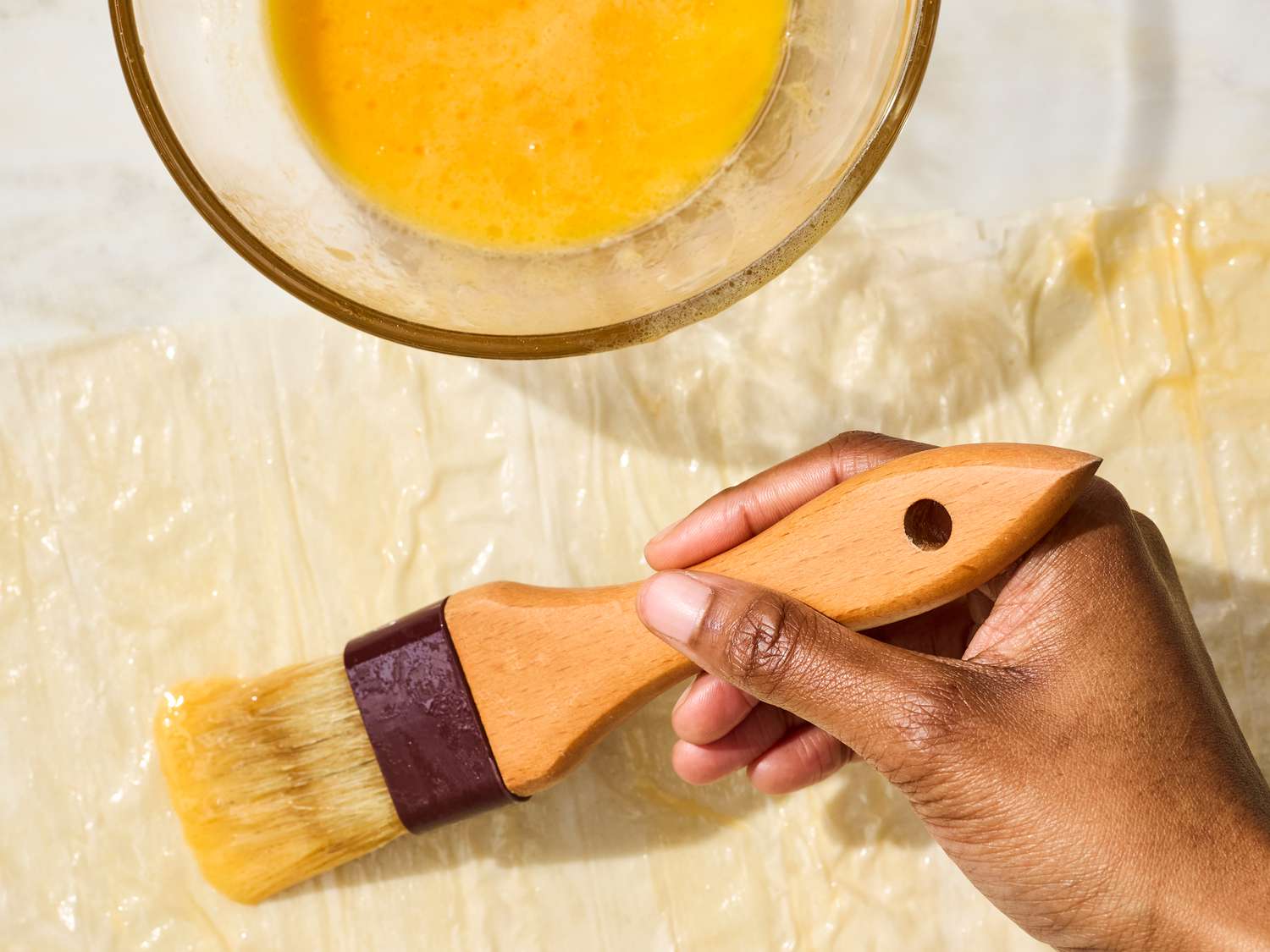 A hand applying egg wash to pastry sheet with a brush, bowl of egg wash in the background