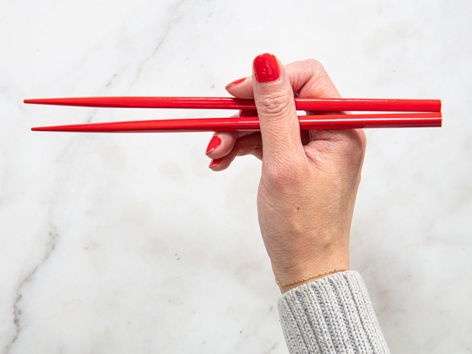 A hand holding red chopsticks on a white background demonstrating how to hold chopsticks