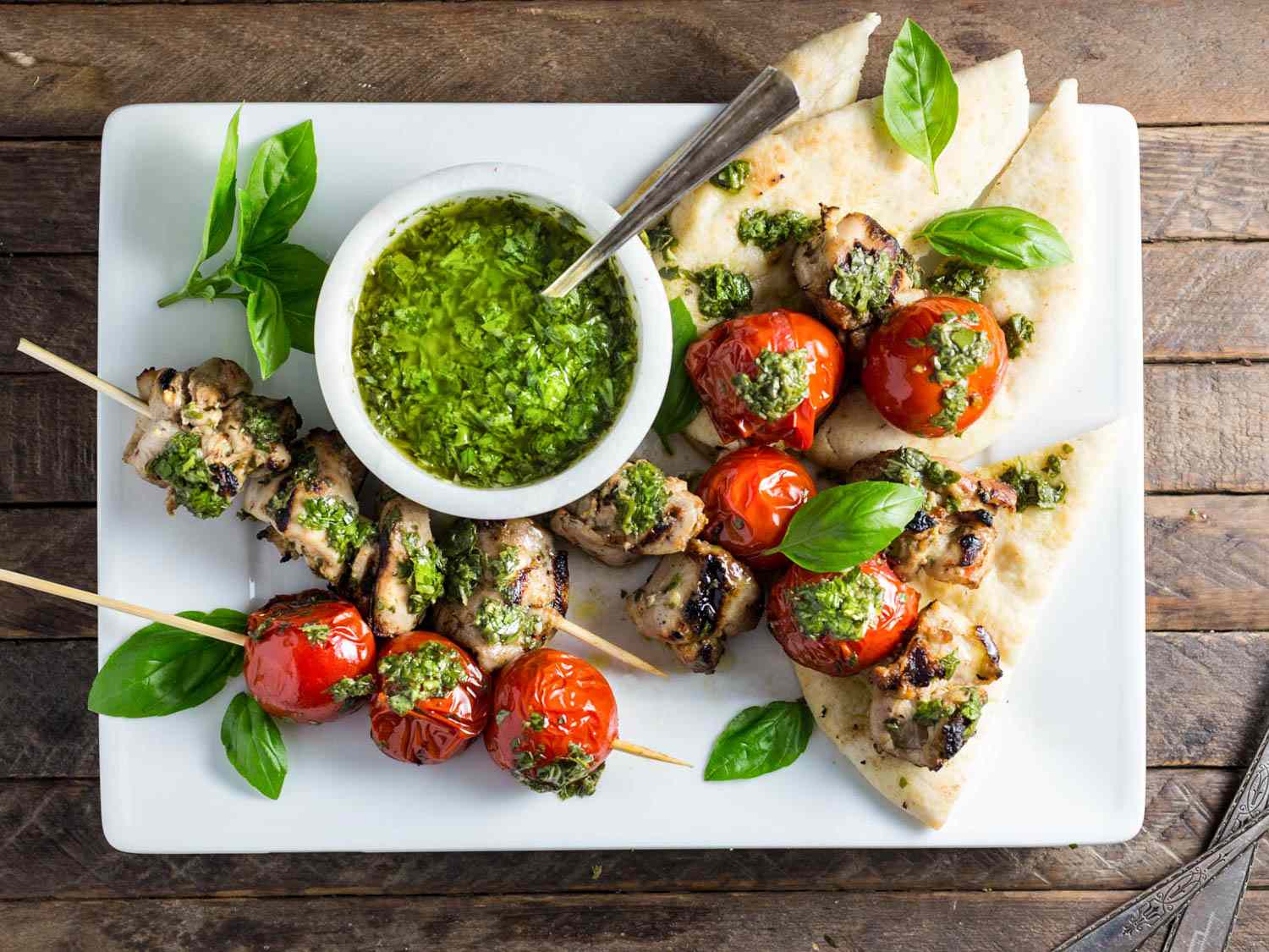 A grilled lemon-garlic chicken kebab and a cherry tomato kebab served on a white rectangular plate with a ramekin of chimichurri and wedges of flatbread.