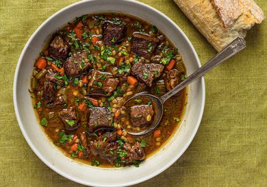 A white porcelain bowl holding beef barley soup. There's a metal spoon in the bowl and the bowl is palced on a green cloth. There is also part of a baguette in the top right corner of the image.
