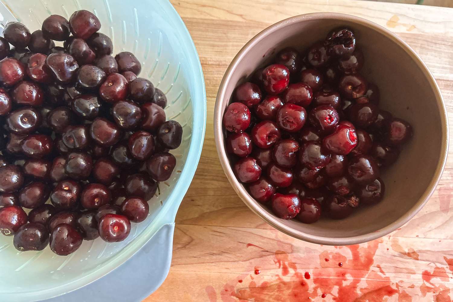A colander full of cherries next to a mixing bowl full of pitted cherries