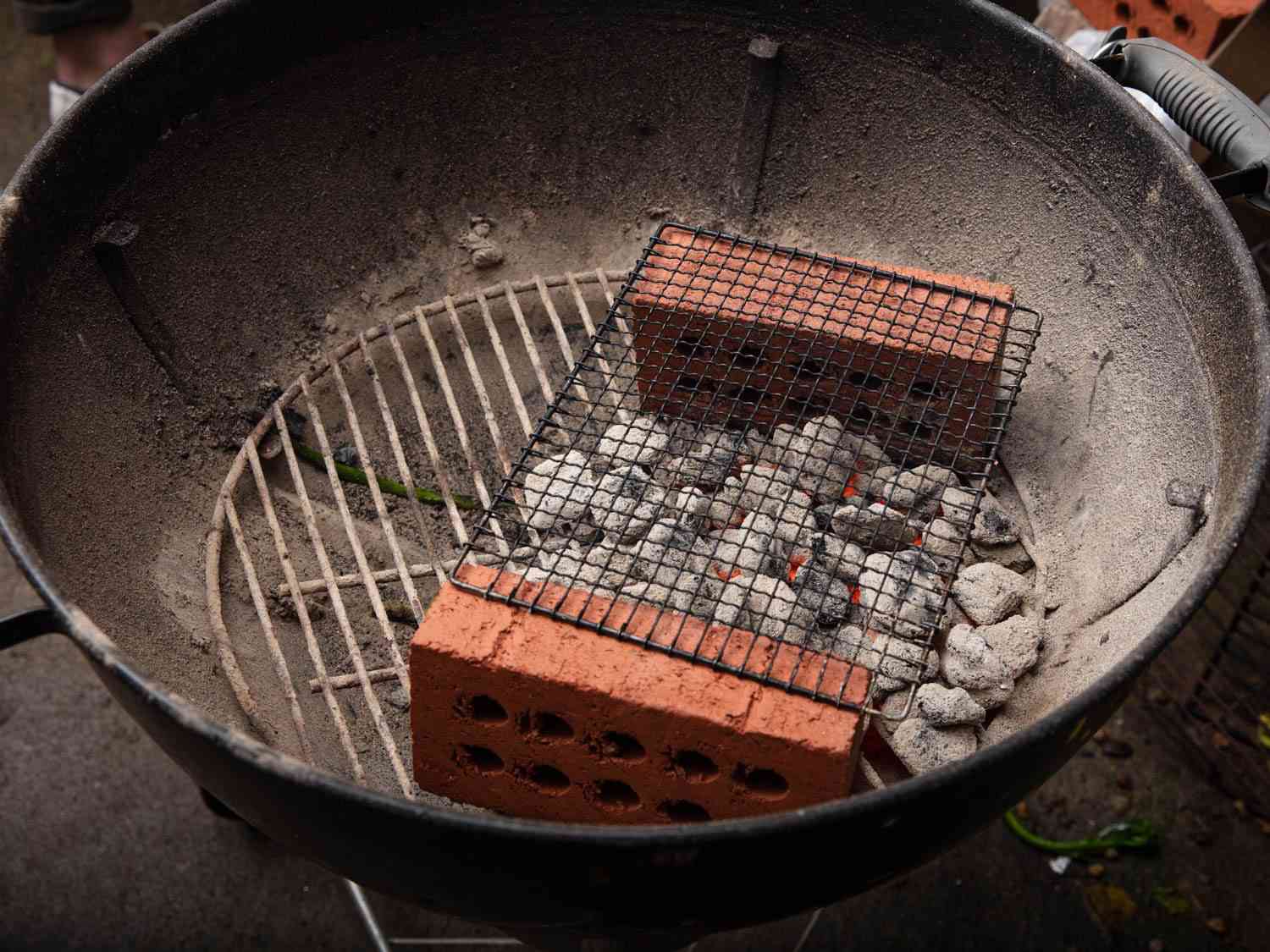 A kettle grill set up for grilling skewers with a mesh wire grilling net set over two bricks, with hot coals positioned between the bricks.