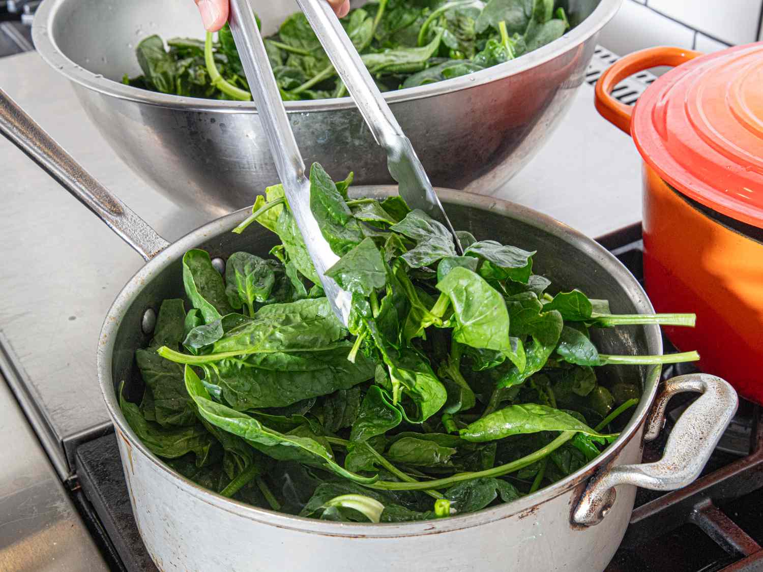 Cooking spinach in a pan using tongs with a red pot in the background